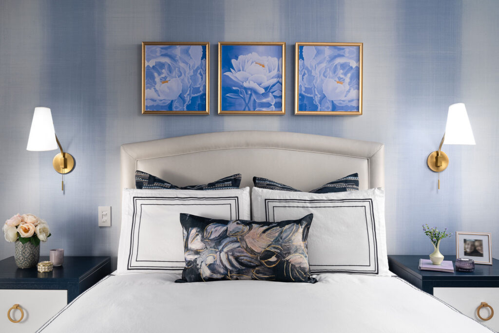 Upholstered faux-leather bed with decorative pillows, navy and white nightstands with gold and white wall sconces above. Above the bed is a trio of peony floral art framed in gold and white mouldings.