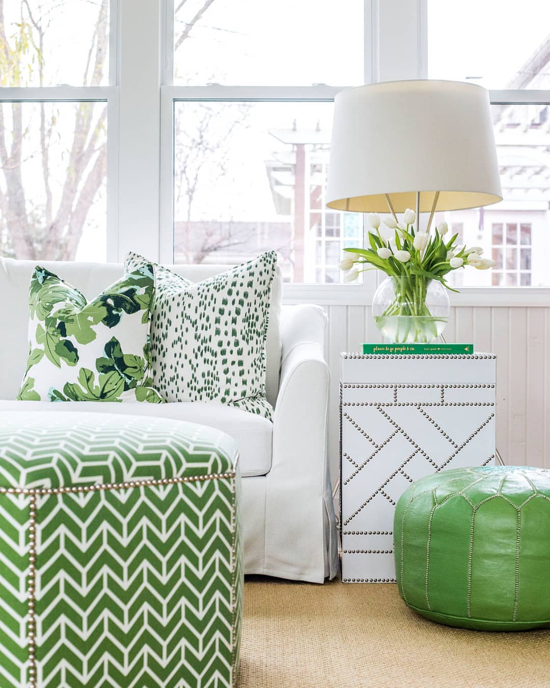 Clean and  contemporary living room in shades of green and white.  White sofa with patterned green ottoman with nailheads, accent pillows is varying green patterned fabrics and a green leather pouf.